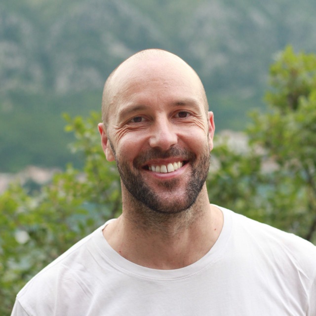Ein lächelnder Mann in einem weißen T-Shirt steht draußen vor einem verschwommenen grünen Hintergrund.