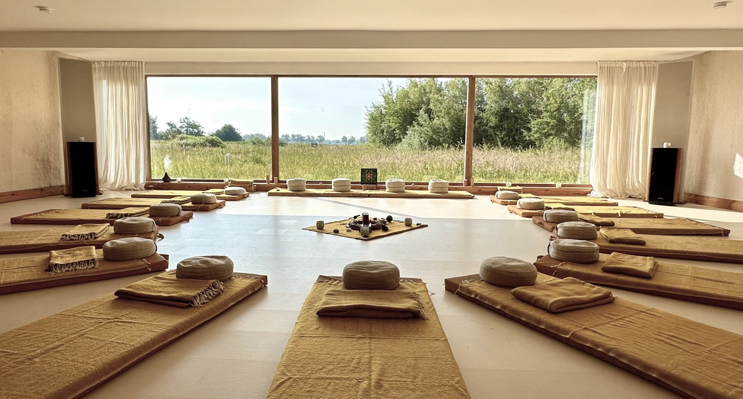 Ein Meditationsraum mit runden Kissen und Matten in einem Kreis angeordnet, mit großen Fenstern, die einen Blick auf eine grüne Landschaft bieten.