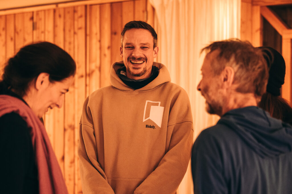 Drei lachende Teilnehmer der Psychedelische Begleitungsausbildung von KULA in warmer Kleidung in einem holzvertäfelten Raum.