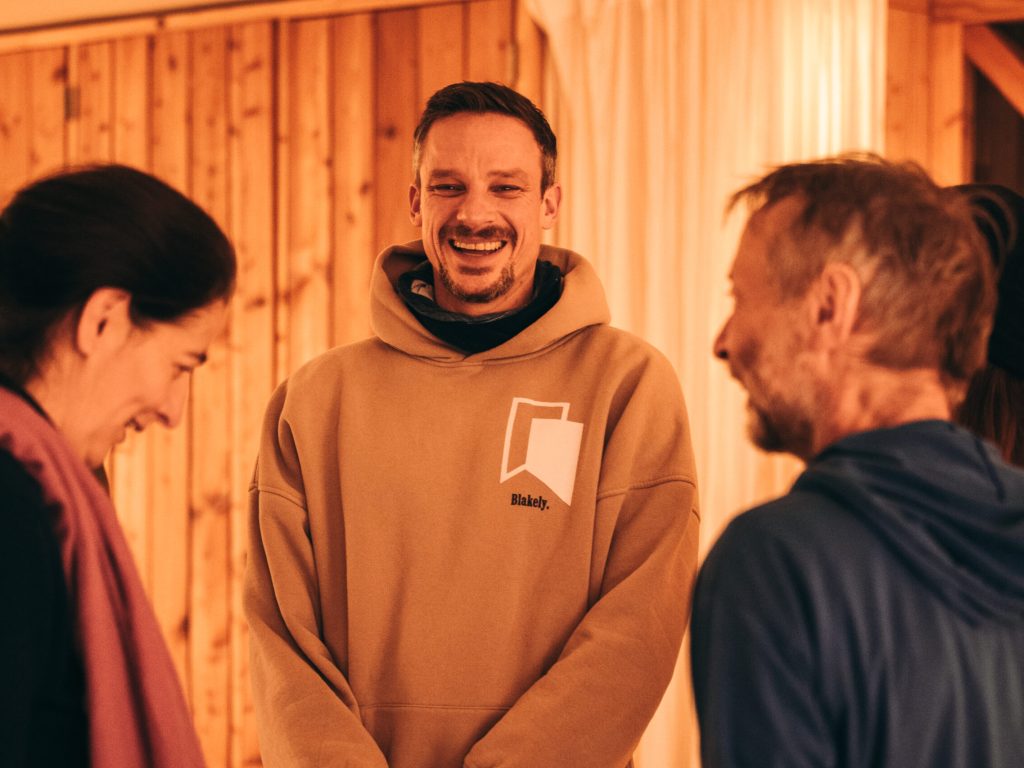 Drei lachende Teilnehmer der Psychedelische Begleitungsausbildung von KULA in warmer Kleidung in einem holzvertäfelten Raum.